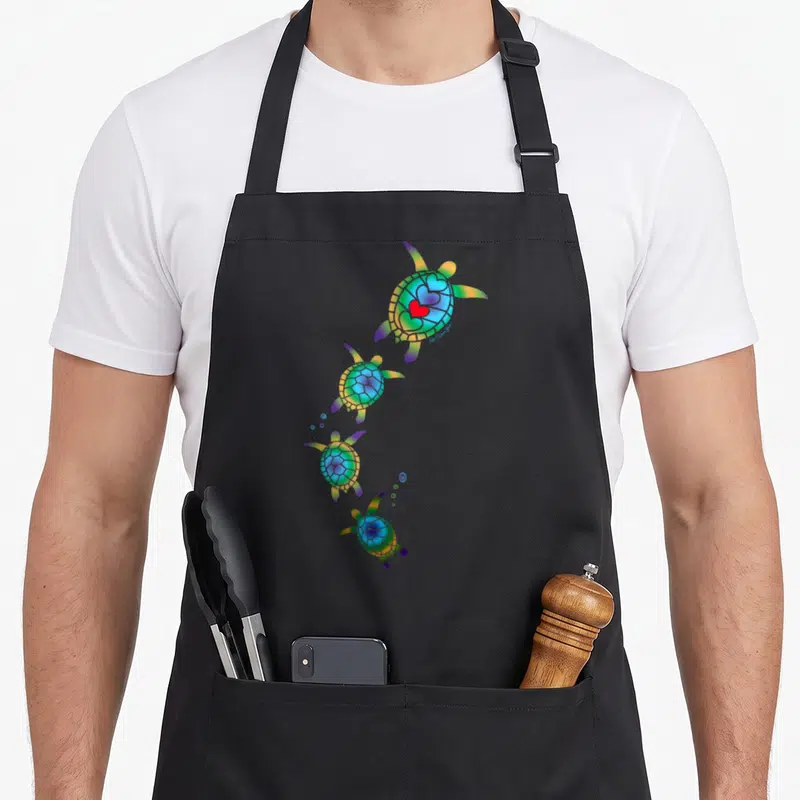 Nautical beautiful apron - Four rainbow sea turtles in a curve with red heart on top shell — shown on a black cotton canvas apron against a white background