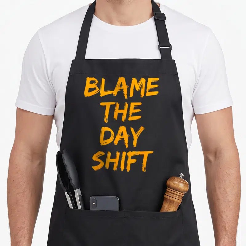 Blame swagazon apron - Orange brush lettering reading BLAME THE DAY SHIFT stacked vertically — shown on a black cotton canvas apron against a white background