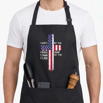 Memorial cross apron - Distressed cross filled with US flag pattern surrounded by white text reading I KNEEL — shown on a black cotton canvas apron against a white background