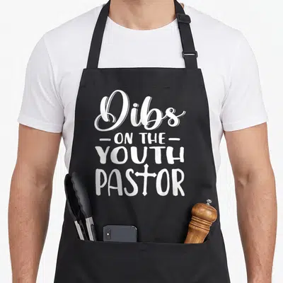 Costume pastor apron - White stacked text reading Dibs on the Youth Pastor with a cross replacing the O in Pastor — shown on a black cotton canvas apron against a white background