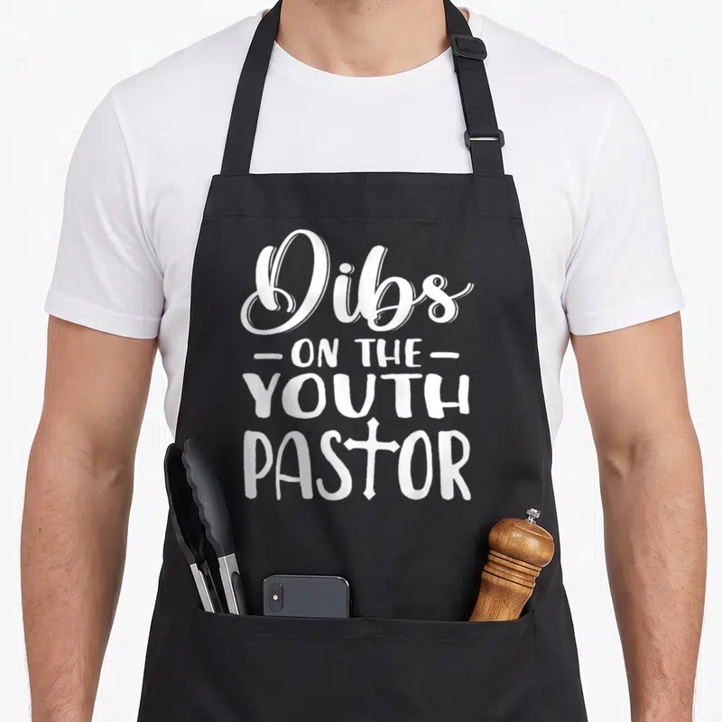 Costume pastor apron - White stacked text reading Dibs on the Youth Pastor with a cross replacing the O in Pastor — shown on a black cotton canvas apron against a white background