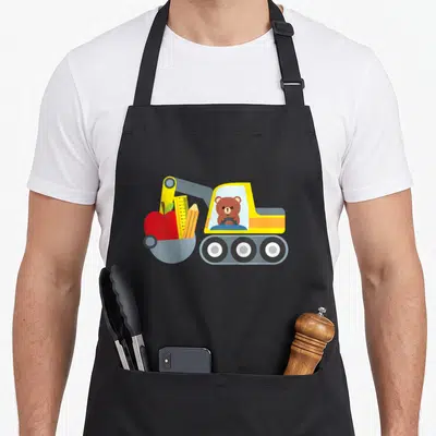 School toddler truck apron - Cartoon excavator with bear driver carrying red apple, yellow ruler, and pencil in bucket — shown on a black cotton canvas apron against a white background