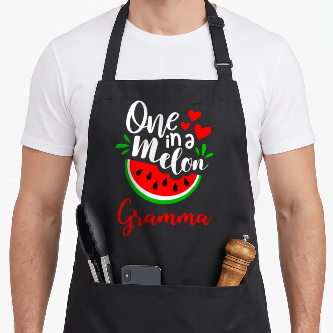 Melon family aprons - One in a Melon Gramma watermelon slice three hearts four green leaves cartoon print — shown on a black cotton canvas apron against a white background