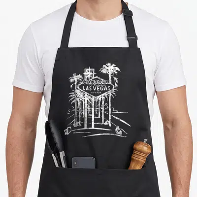 Las famous aprons - White distressed Welcome to Fabulous Las Vegas sign with two palm trees and star — shown on a black cotton canvas apron against a white background