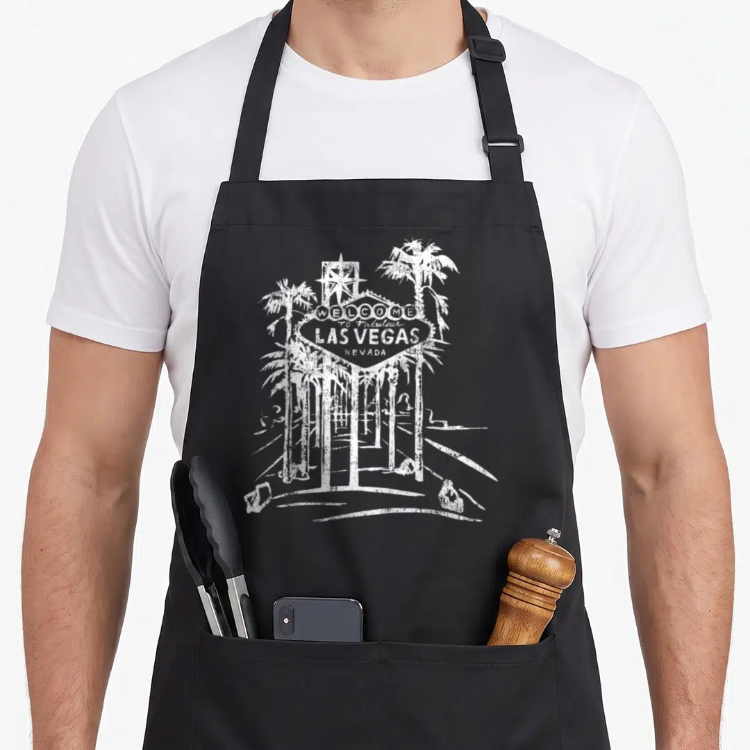 Las famous aprons - White distressed Welcome to Fabulous Las Vegas sign with two palm trees and star — shown on a black cotton canvas apron against a white background