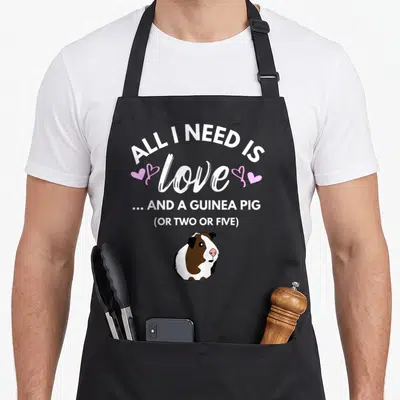 Funny pig i love my guinea apron - Stacked text reading All I Need Is Love And A Guinea Pig Or Two Or Five above — shown on a black cotton canvas apron against a white background