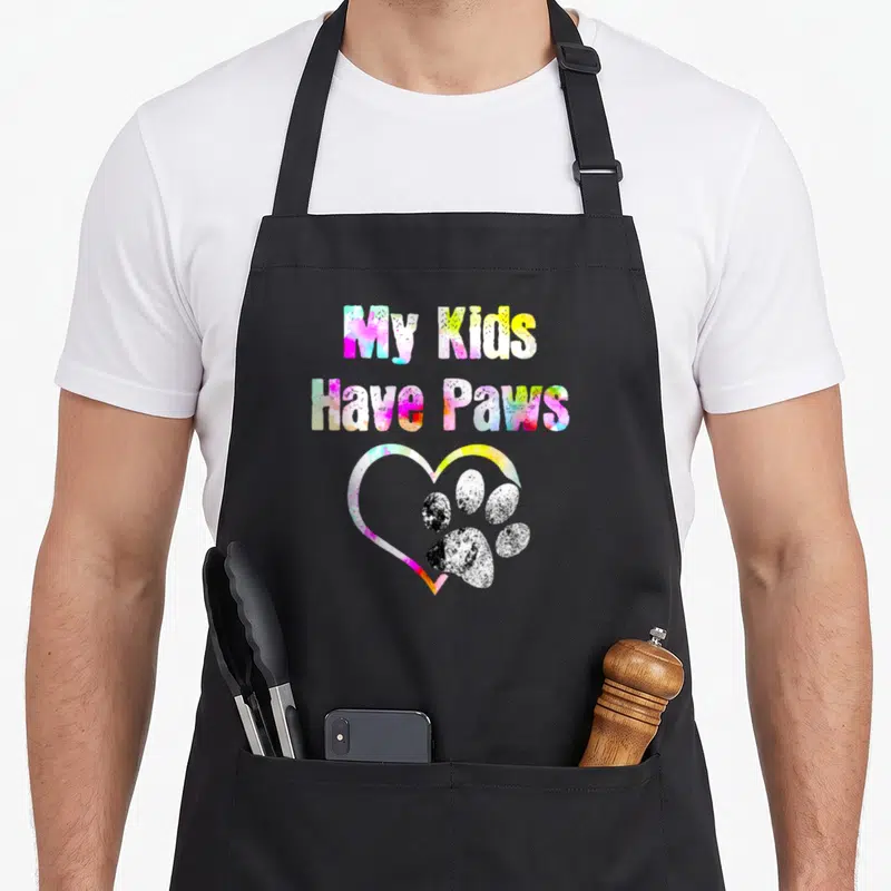 Funny paw cat apron - My Kids Have Paws text above multicolored heart with black and white paw print — shown on a black cotton canvas apron against a white background
