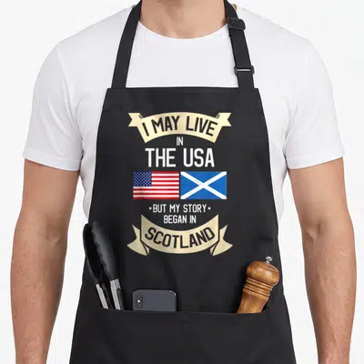Scotland american flag apron - Stacked text with USA and Scottish flags on beige ribbons — shown on a black cotton canvas apron against a white background