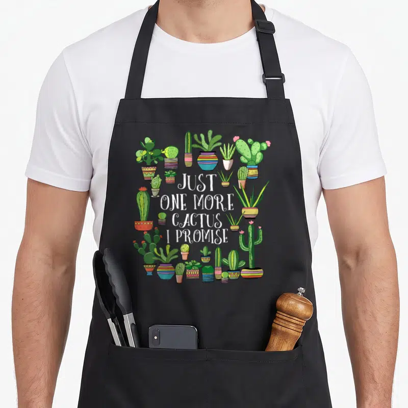 Promise cactus apron - Just one more cactus I promise text surrounded by colorful potted cacti in cartoon style — shown on a black cotton canvas apron against a white background
