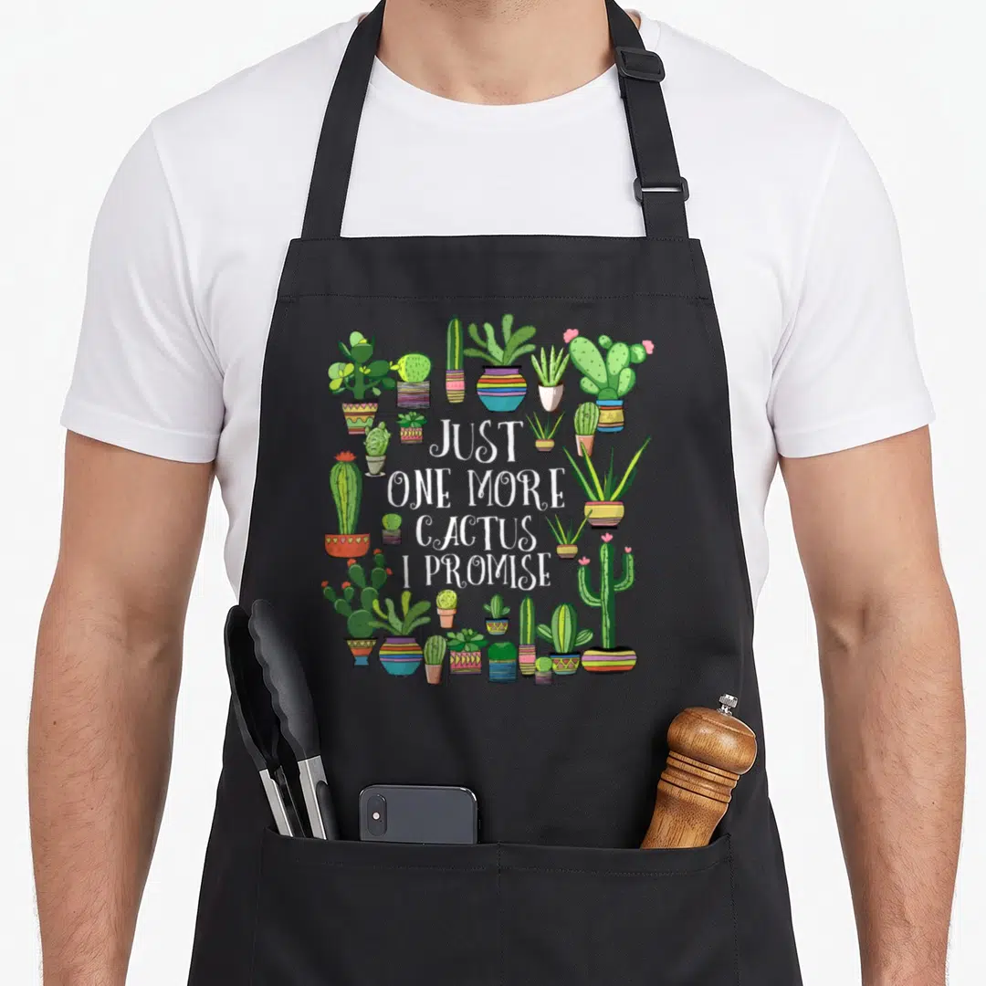 Promise cactus apron - Just one more cactus I promise text surrounded by colorful potted cacti in cartoon style — shown on a black cotton canvas apron against a white background