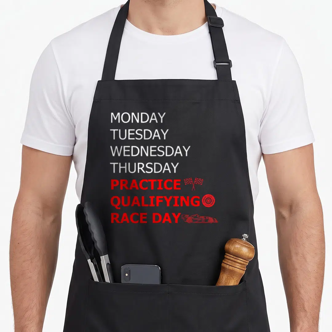 Monday training apron - Stacked text MONDAY through RACE DAY with red flags, tire, and car icons on white and red type — shown on a black cotton canvas apron against a white background