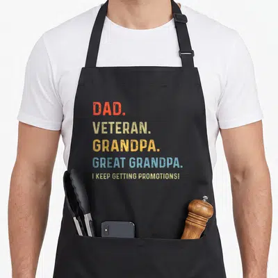 Funny veteran father day apron - Stacked DAD VETERAN GRANDPA GREAT GRANDPA text in red beige yellow blue colors — shown on a black cotton canvas apron against a white background