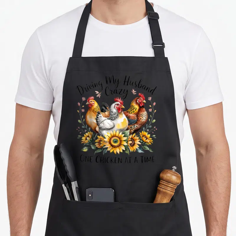 Funny husband driving aprons - Three chickens and sunflowers with Driving My Husband Crazy text — shown on a black cotton canvas apron against a white background