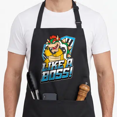 Bold boss apron - Cartoon Bowser character with gradient LIKE A BOSS text and blue shield background — shown on a black cotton canvas apron against a white background