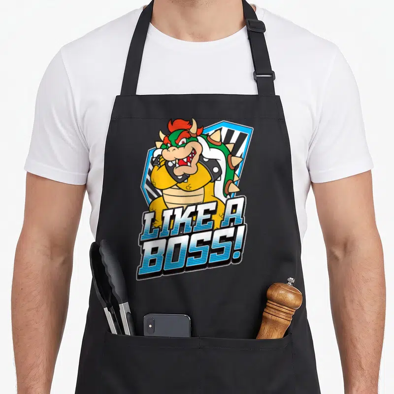 Bold boss apron - Cartoon Bowser character with gradient LIKE A BOSS text and blue shield background — shown on a black cotton canvas apron against a white background