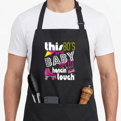 80s hanging apron - Stacked text reading this 80s baby still hangin touch with roller skate cassette and sunglasses — shown on a black cotton canvas apron against a white background