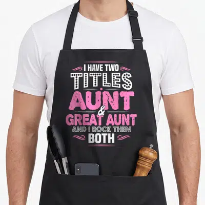 Aunt title apron - Pink and white typography reading I Have Two Titles Aunt and Great Aunt flanked by swirls — shown on a black cotton canvas apron against a white background