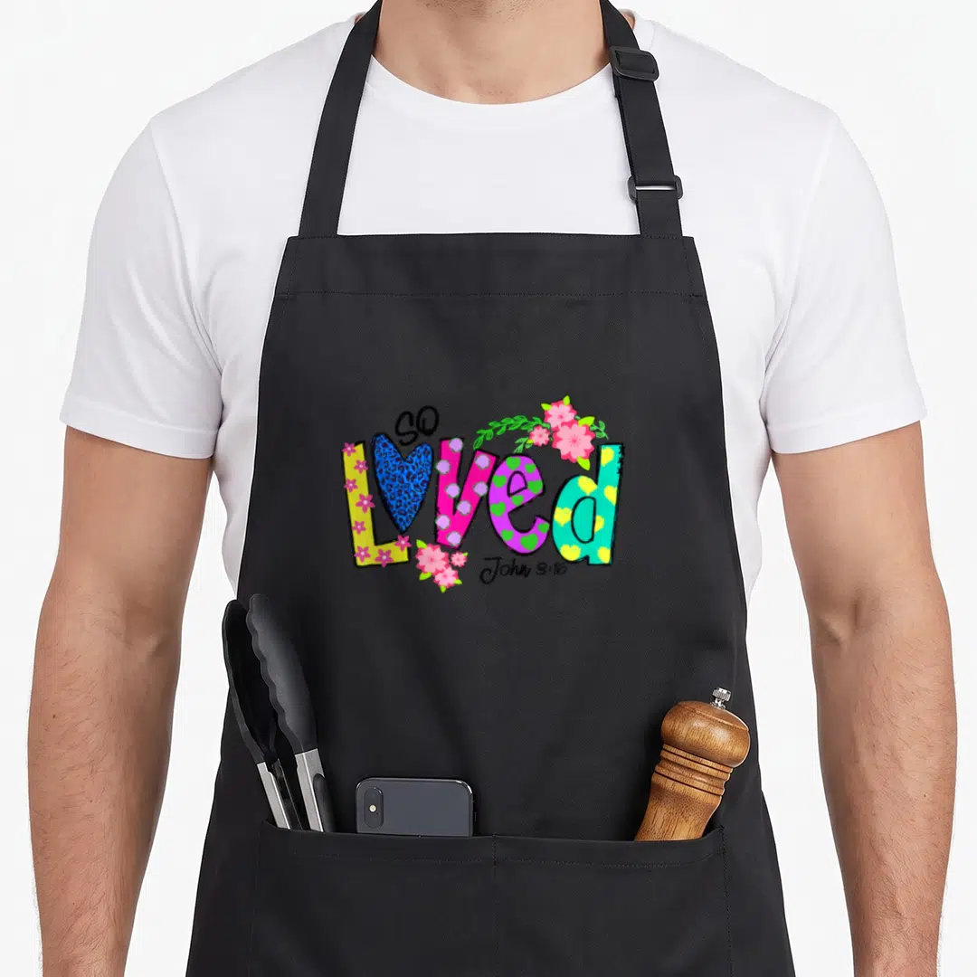 Loved polka dot apron - Cartoon letters so loved John 3:16 with heart and flowers in pink teal yellow blue green — shown on a black cotton canvas apron against a white background