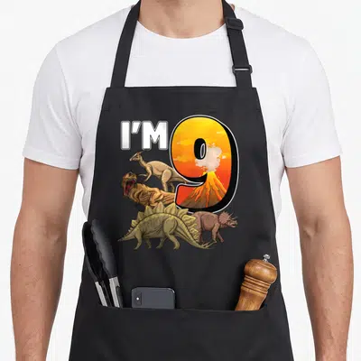Birthday 9th dinosaur apron - I'M 9 text next to volcano-filled number 9 surrounded by four cartoon dinosaurs — shown on a black cotton canvas apron against a white background