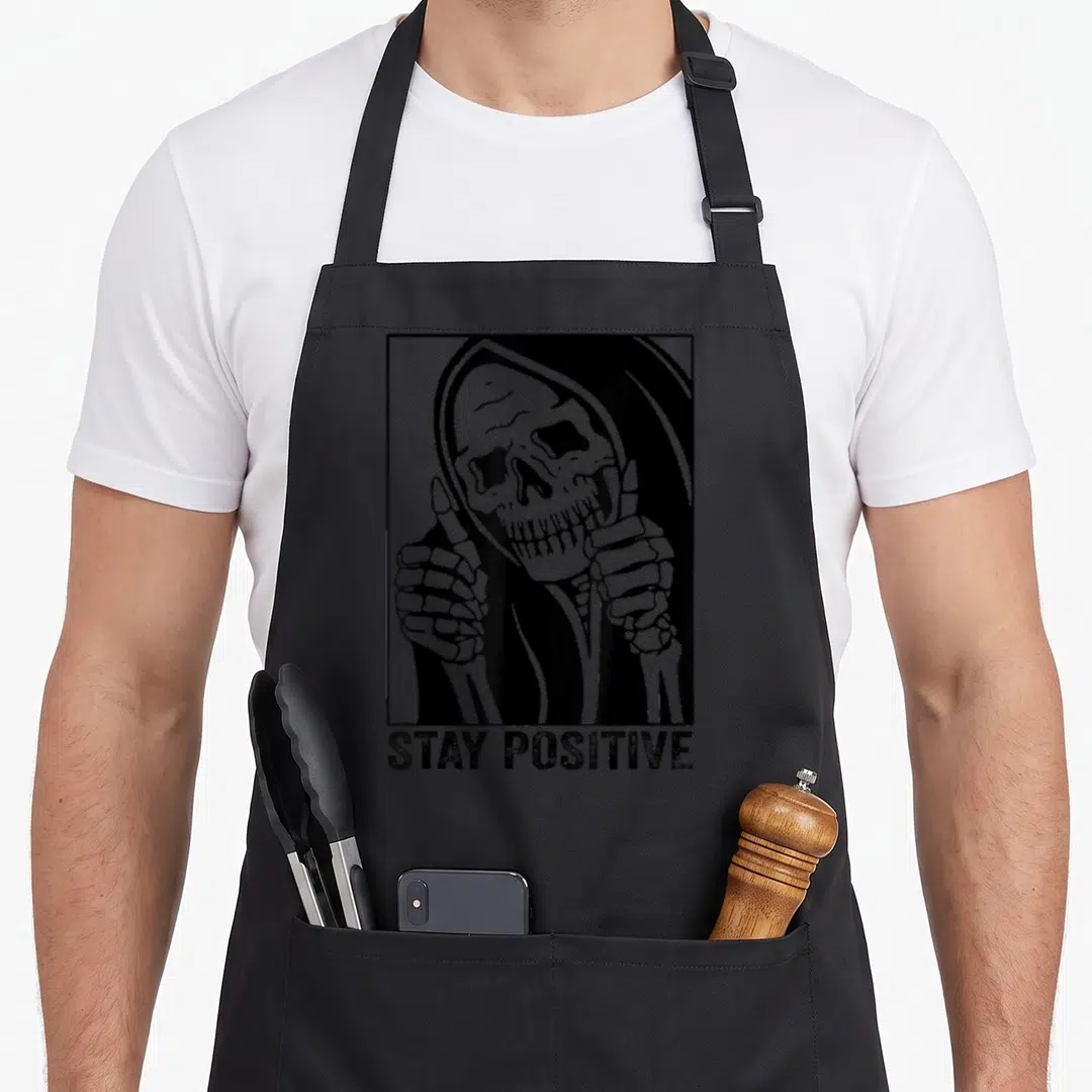 Stay skeleton apron - Cartoon Grim Reaper in square frame giving two thumbs up above STAY POSITIVE text — shown on a black cotton canvas apron against a white background