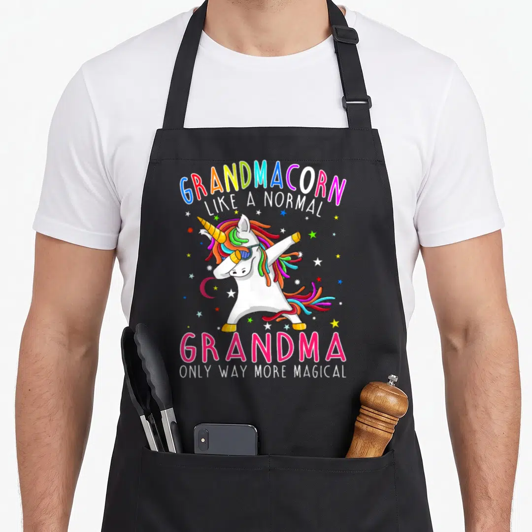 Grandmacorn grandma apron - Dabbing unicorn with rainbow mane and sunglasses surrounded by stars and crescent moon — shown on a black cotton canvas apron against a white background
