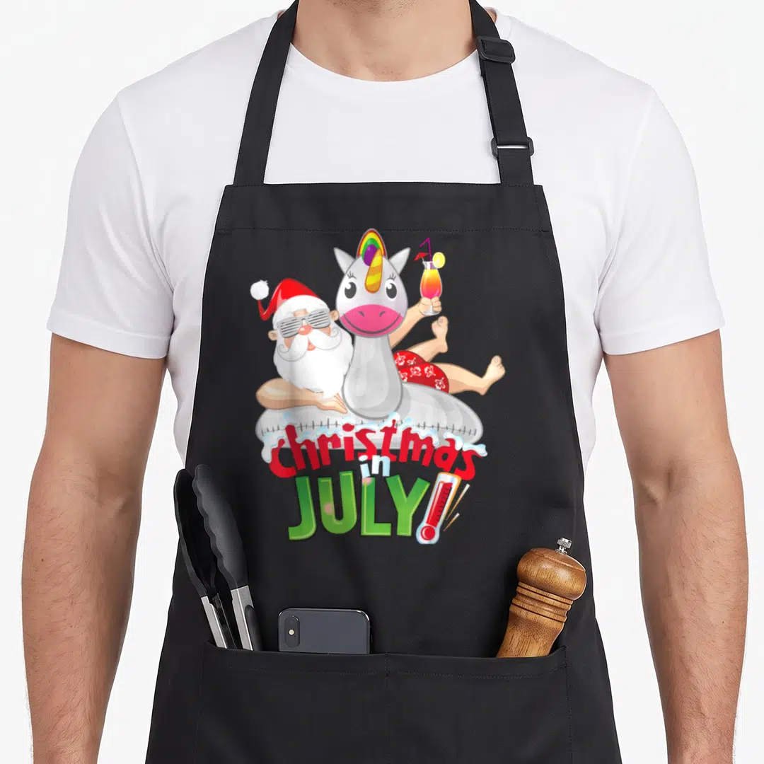 Float christmas apron - Santa in unicorn float holding cocktail with Christmas in July text and thermometer — shown on a black cotton canvas apron against a white background