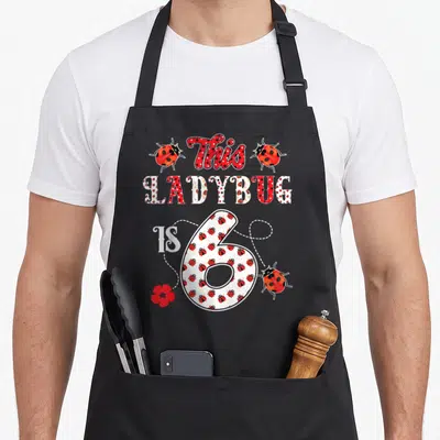 6th ladybug apron - This LADYBUG IS 6 text with red ladybugs, flower, and dotted lines — shown on a black cotton canvas apron against a white background