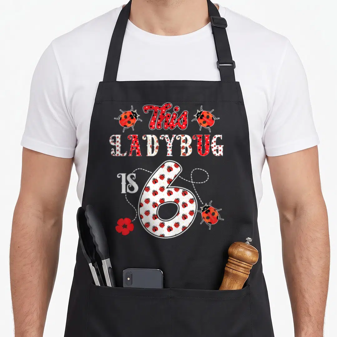 6th ladybug apron - This LADYBUG IS 6 text with red ladybugs, flower, and dotted lines — shown on a black cotton canvas apron against a white background