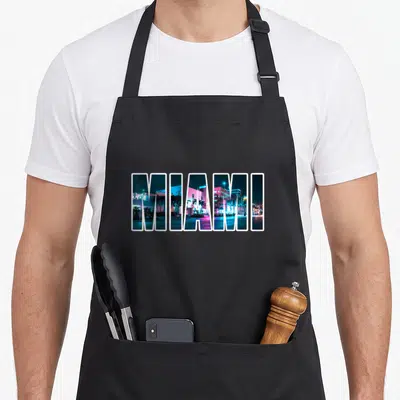 Miami urban apron - MIAMI text filled with blue and pink night cityscape photo — shown on a black cotton canvas apron against a white background