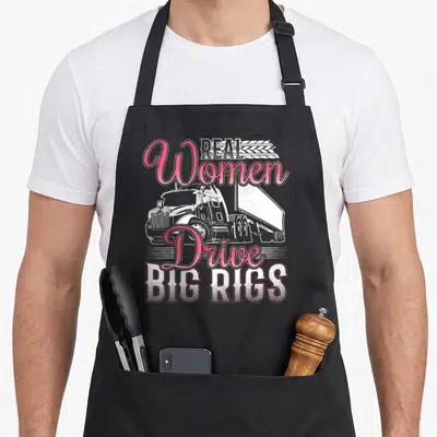 Drive rig apron - Real Women Drive Big Rigs text surrounding a white semi-truck illustration in retro style — shown on a black cotton canvas apron against a white background