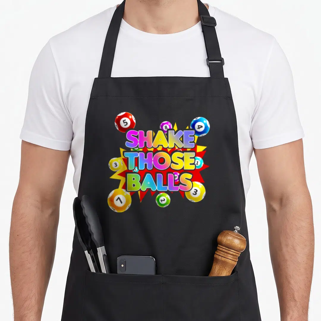 Shake bingo aprons - Stacked Shake Those Balls text over red yellow explosion surrounded by numbered lottery balls — shown on a black cotton canvas apron against a white background