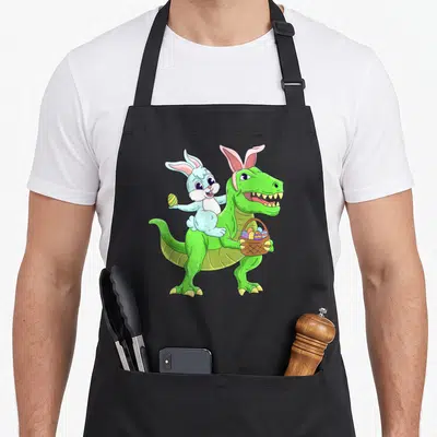 Riding easter bunny aprons - Light blue rabbit riding green dinosaur holding basket of colorful eggs — shown on a black cotton canvas apron against a white background