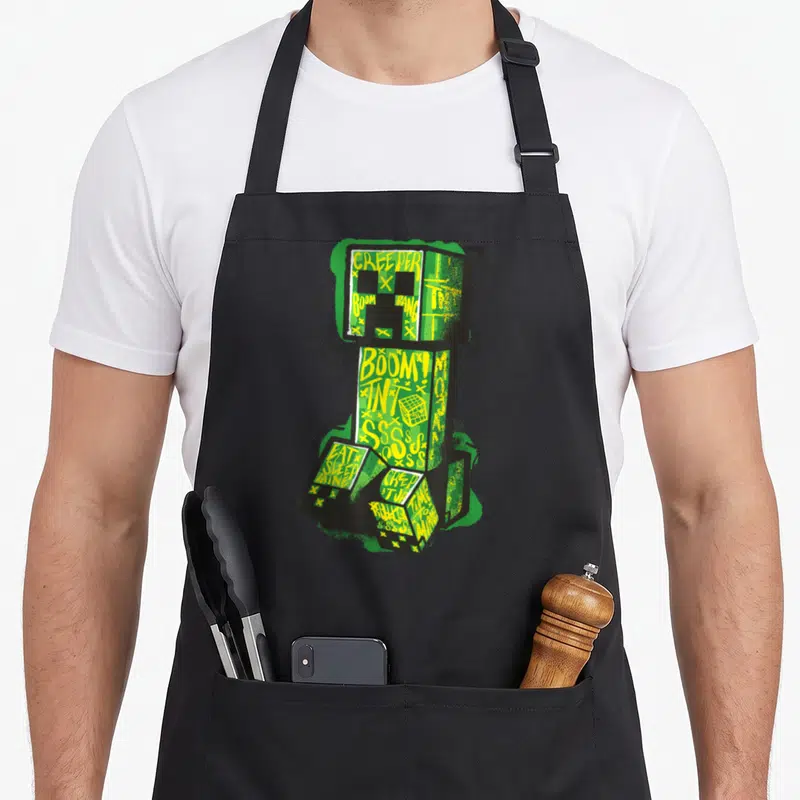 Creeper street apron - Green creeper figure with yellow graffiti text BOOM TNT SSSS and spray paint effects — shown on a black cotton canvas apron against a white background