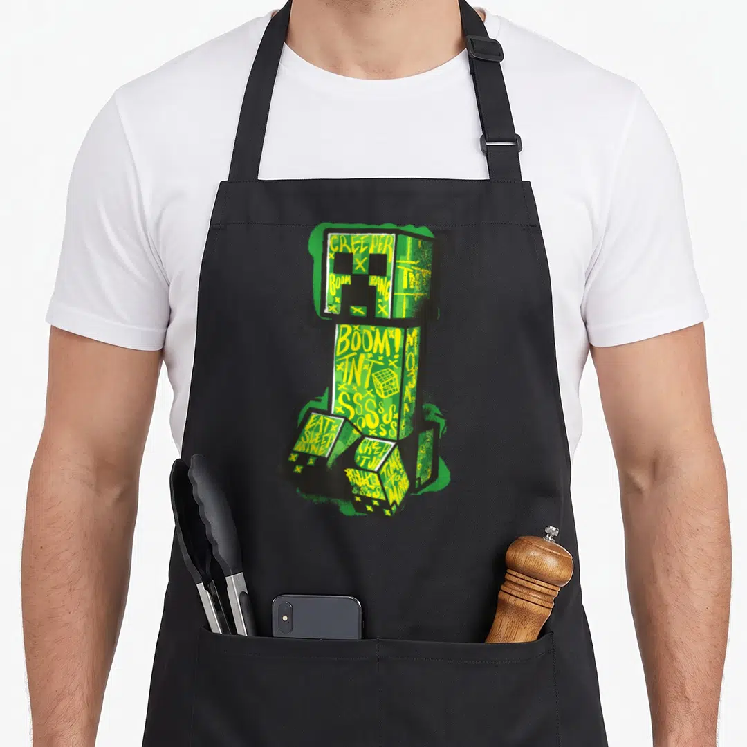 Creeper street apron - Green creeper figure with yellow graffiti text BOOM TNT SSSS and spray paint effects — shown on a black cotton canvas apron against a white background