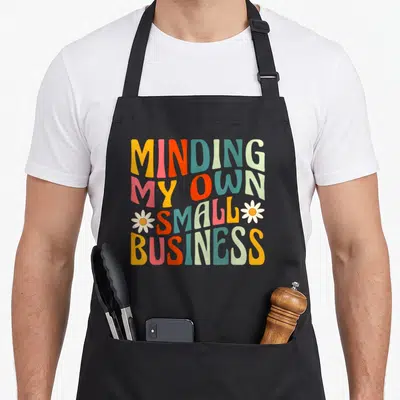 Minding small business aprons - Retro colorful text reading MINDING MY OWN SMALL BUSINESS flanked by two white daisies — shown on a black cotton canvas apron against a white background
