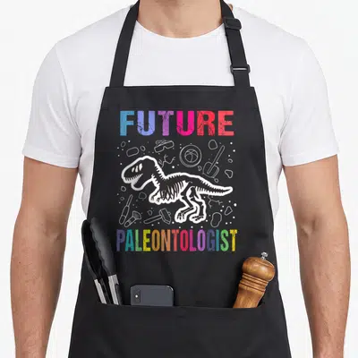 Future paleontologist dinosaur apron - Retro dinosaur skeleton with FUTURE PALEONTOLOGIST text and white tool outlines — shown on a black cotton canvas apron against a white background