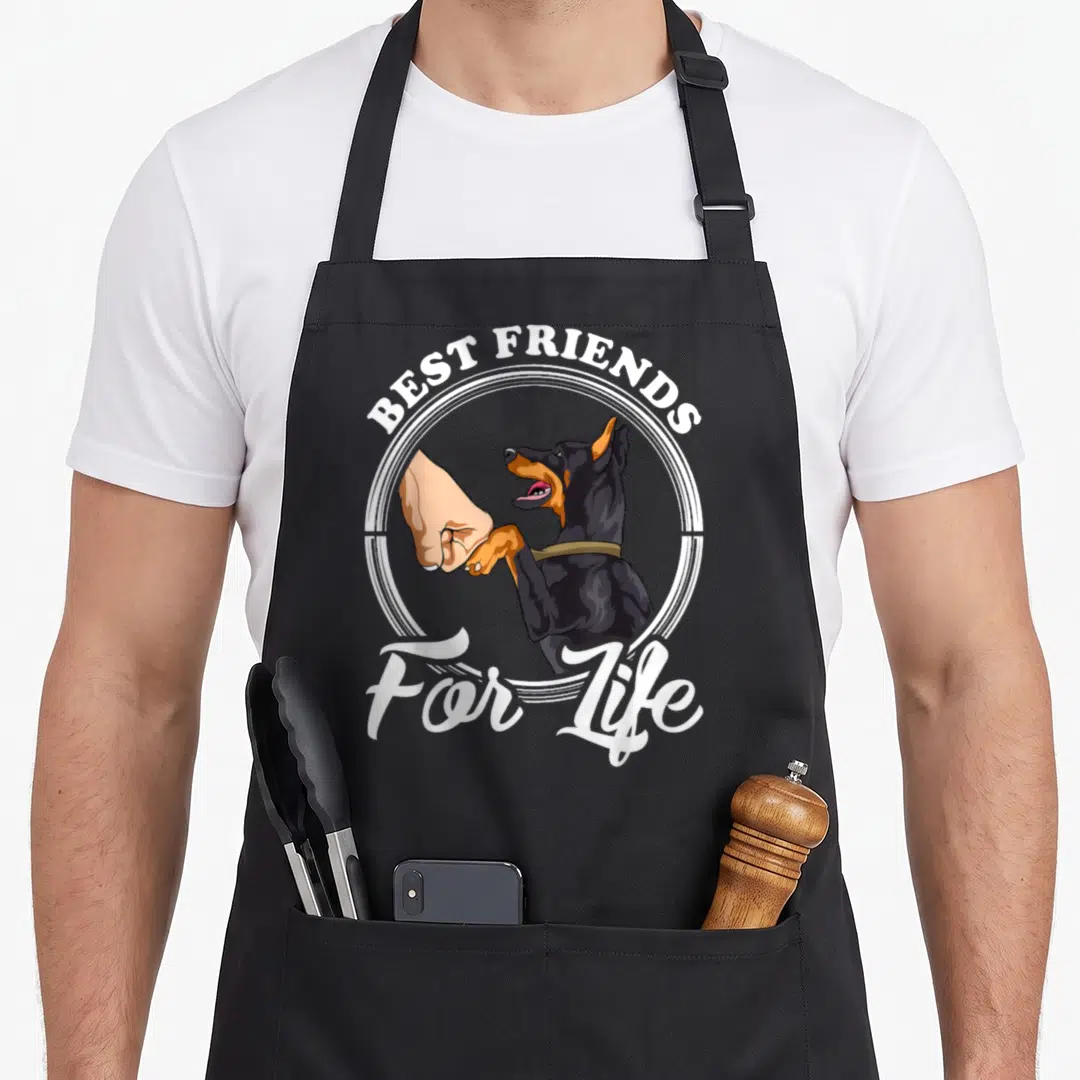 Funny pinscher doberman apron - Cartoon doberman fist-bumping a human hand with best friends for life text in a white — shown on a black cotton canvas apron against a white background