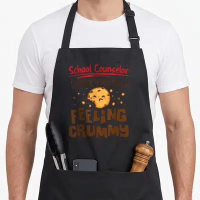 Elementary school counselor apron - Sad cartoon cookie with bite mark and text School Counselor Feeling Crummy — shown on a black cotton canvas apron against a white background