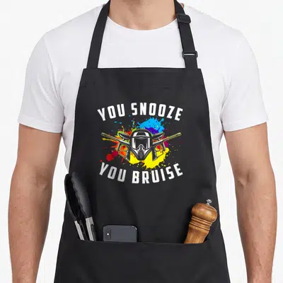 Youth paintball apron - YOU SNOOZE YOU BRUISE text around mask and crossed markers with paint splatters — shown on a black cotton canvas apron against a white background