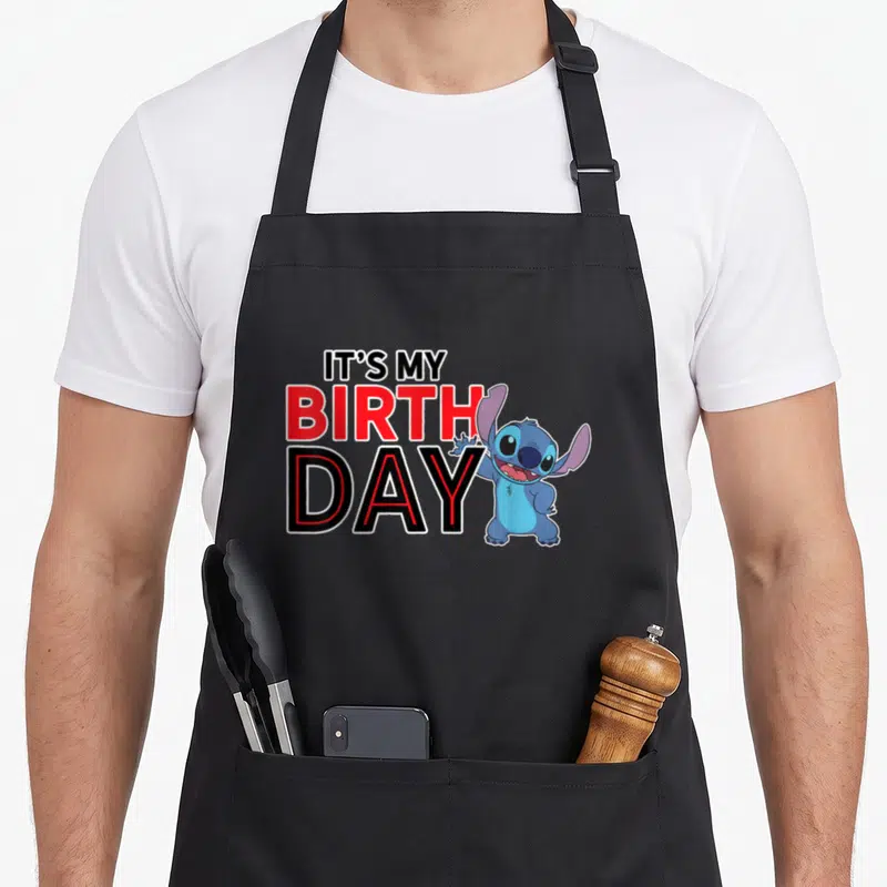 Cute age birthday apron - Waving blue Stitch next to stacked IT'S MY BIRTH DAY text in red and black — shown on a black cotton canvas apron against a white background
