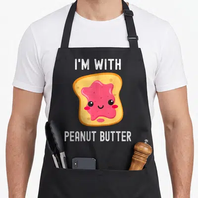 His butter apron - Cartoon toast with smiling pink jam face and white text reading I'M WITH PEANUT BUTTER — shown on a black cotton canvas apron against a white background
