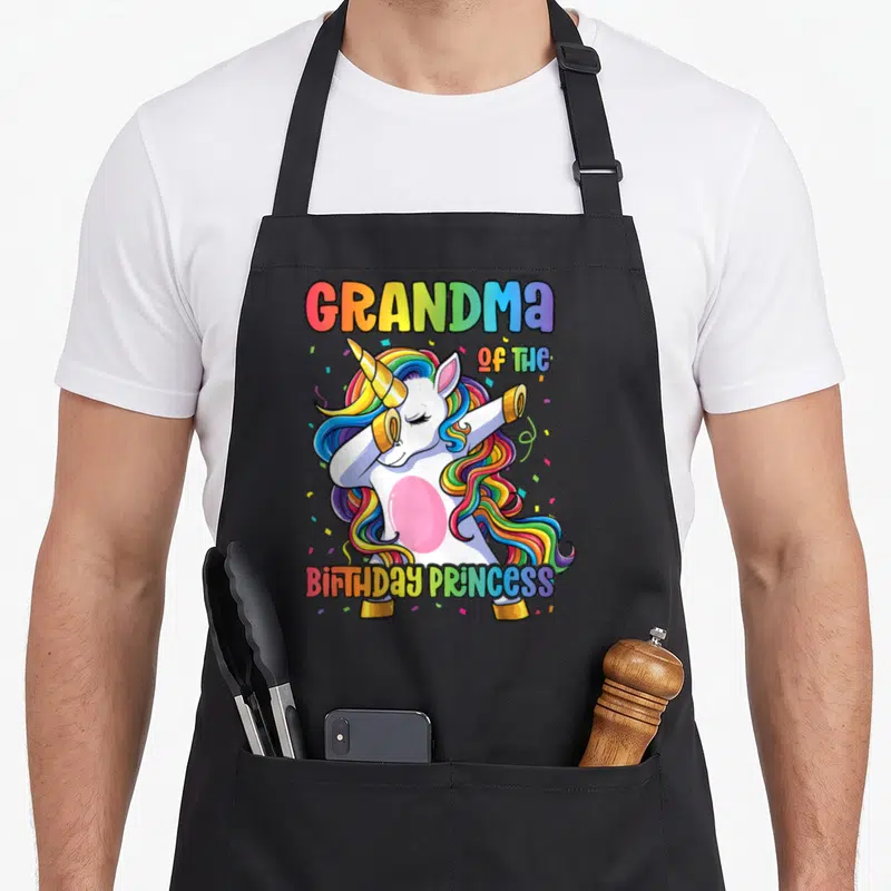 Princess dabbing grandma in apron - Cartoon unicorn dabbing with rainbow mane and GRANDMA OF THE BIRTHDAY PRINCESS text — shown on a black cotton canvas apron against a white background