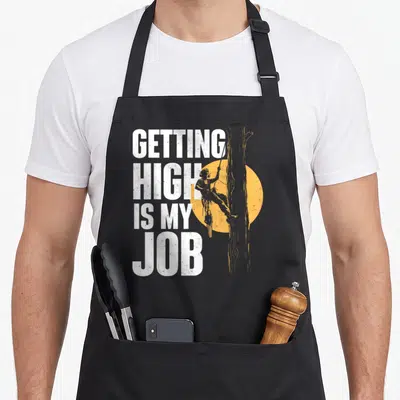 Getting surgeon apron - White stacked text GETTING HIGH IS MY JOB next to orange arborist climbing tree silhouette — shown on a black cotton canvas apron against a white background