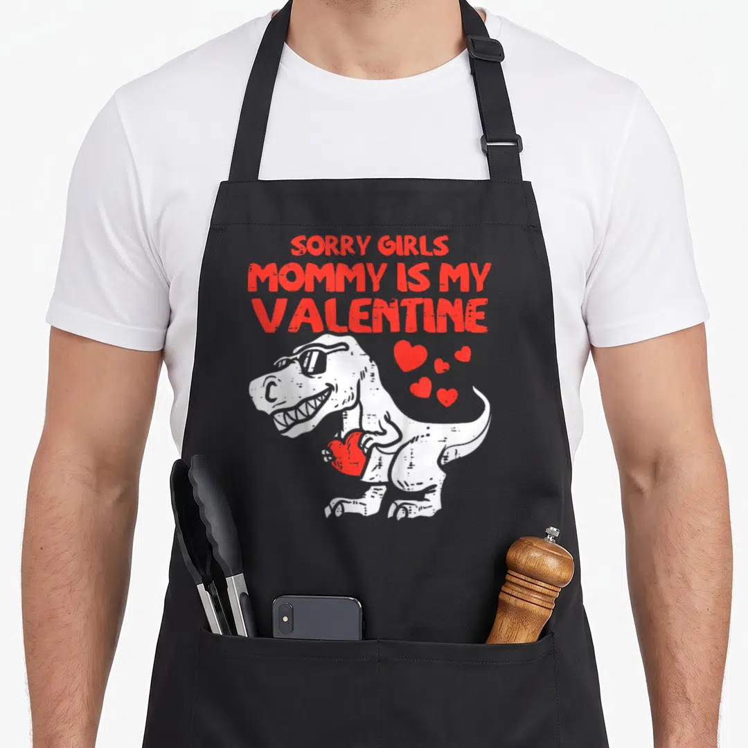 Funny sorry apron - Red text sorry girls mommy is my valentine above white dino holding heart — shown on a black cotton canvas apron against a white background