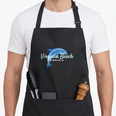 Beach virginia apron - Blue cartoon dolphin with Virginia Beach text overlay and two front pockets — shown on a black cotton canvas apron against a white background