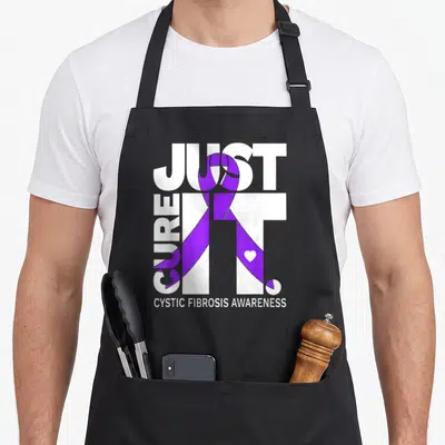 Fibrosis ribbon apron - White text Just Cure It with purple ribbon and heart on black bib — shown on a black cotton canvas apron against a white background