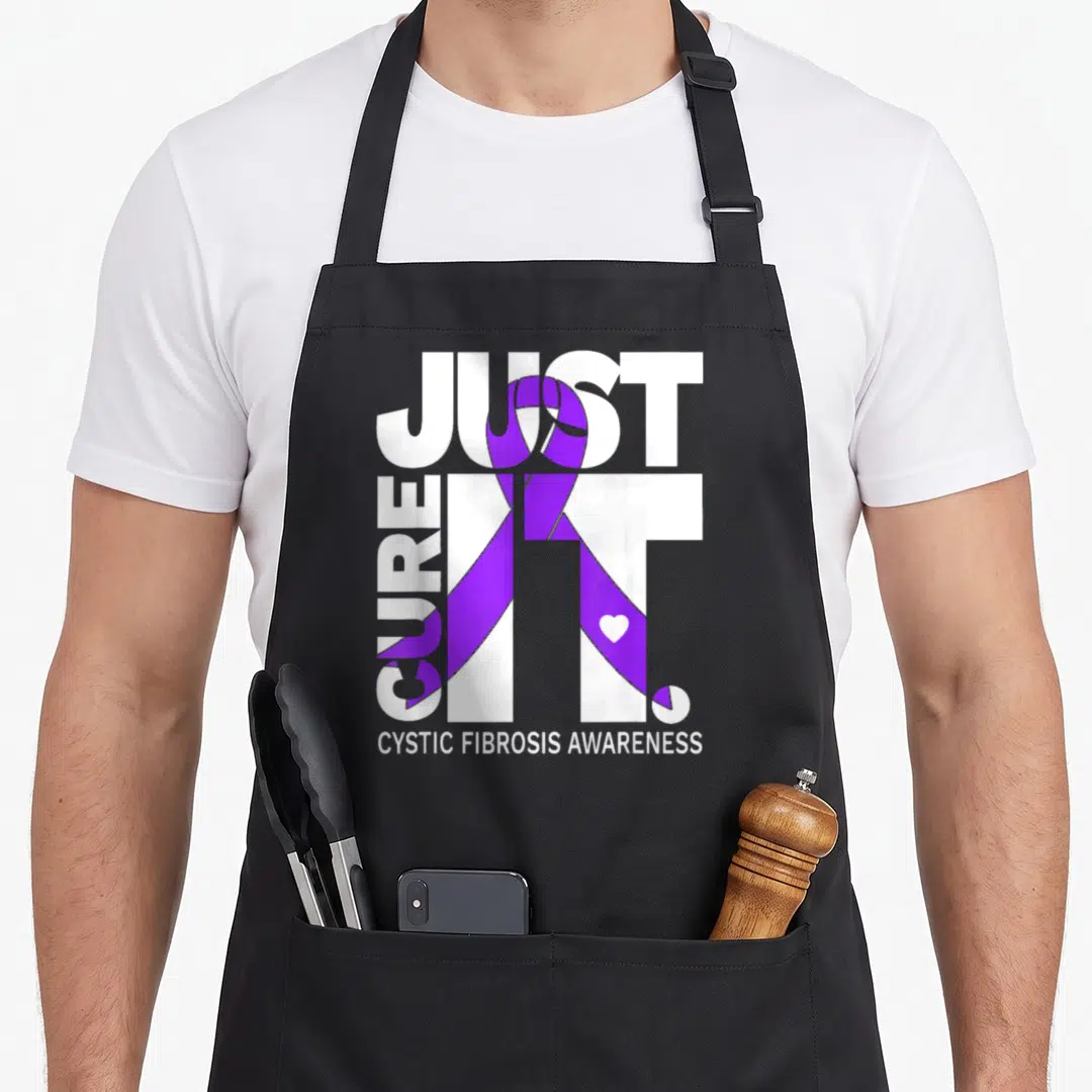 Fibrosis ribbon apron - White text Just Cure It with purple ribbon and heart on black bib — shown on a black cotton canvas apron against a white background