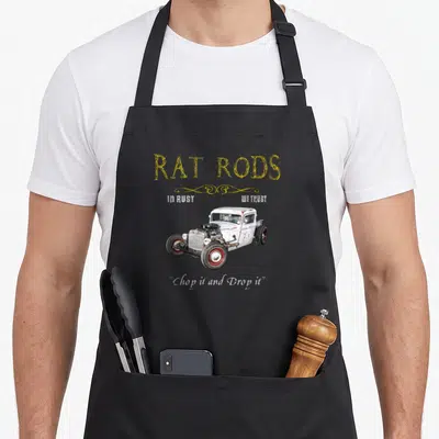 Rod crazy rat man apron - Rat rod car with yellow swirls and red wheels, white text reading In Rust We Trust — shown on a black cotton canvas apron against a white background