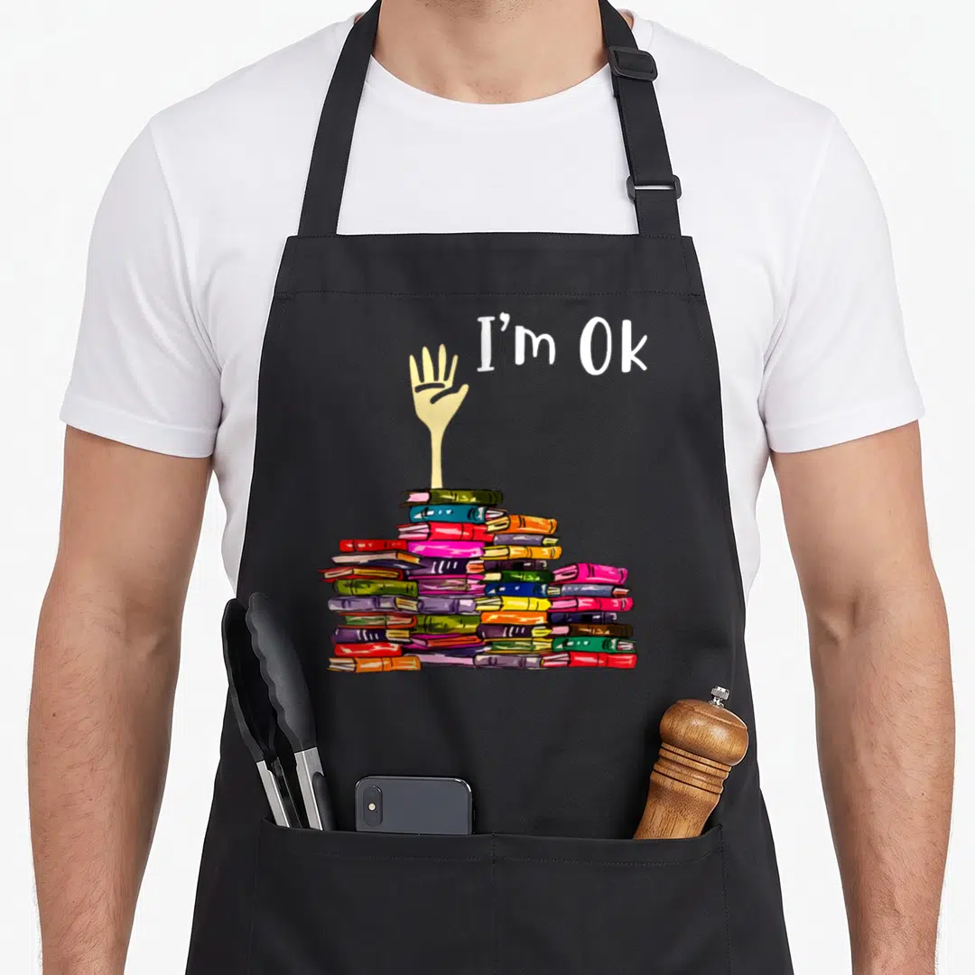 Book teacher apron - I'm Ok text next to hand rising from stack of red blue green yellow white books — shown on a black cotton canvas apron against a white background