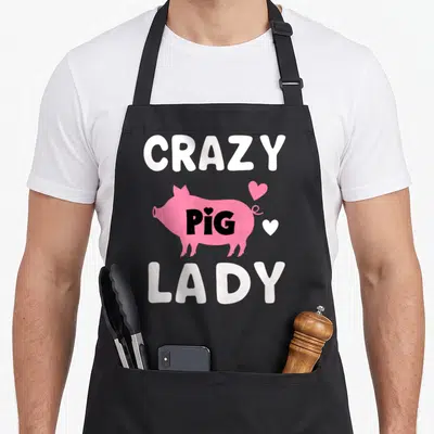 Pig farmer apron for women - CRAZY PIG LADY text surrounding a pink pig with two hearts in cartoon style — shown on a black cotton canvas apron against a white background